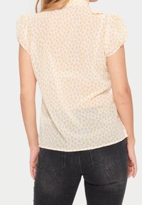 Blouse à manches courtes, transparente, de couleur jaune clair avec des taches orange. Encolure arrondie, épaules légèrement froncées et coupe décontractée.