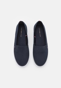 Zapatillas de lona azul marino sin cordones con una suela de goma blanca, que cuentan con un logo en la planta y acentos en rojo, blanco y azul en el talón.