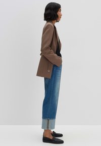 Blazer marrone sopra una maglietta nera, abbinato a jeans a gamba larga blu chiaro con orli e scarpe slip-on nere con un motivo testurizzato.