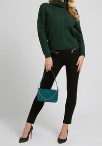 Pull vert en maille torsadée à col roulé, pantalon noir ajusté, et sac à main turquoise avec bandoulière chaîne. Escarpins noirs à bout pointu.