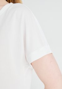 Chemise blanche à manches courtes avec un décolleté en V, fabriquée en tissu doux. Présente des poignets retroussés et une coupe décontractée, mettant en avant une texture et un design doux.