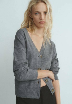 Cardigan en laine grise avec un col en V, des manches longues et un ourlet côtelé. Il présente quatre boutons marron sur le devant et une texture lisse.