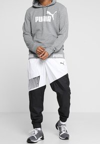 Sudadera gris con el logo blanco de "PUMA", combinada con pantalones de chándal ajustados en blanco y negro que presentan acentos de malla y un patrón geométrico audaz. Zapatillas negras.