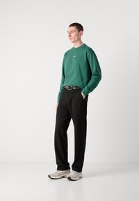Sudadera verde con cuello redondo y logo, combinada con pantalones negros y zapatillas blancas. Tejido suave, corte relajado y diseño minimalista.