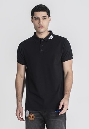 Camisa polo negra con cuello, que presenta tres botones. Llevada con jeans, adornados con parches coloridos en los bolsillos.