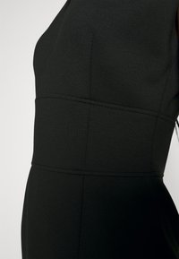 Musta muotoileva mekko, jossa on jämäkkää ja sileää kangasta. Sisältää pystysuoria saumoja ja hienovaraisen peplum-efektin helman kohdalla.