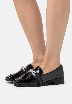 Mocassins en cuir verni noir avec un talon plat, présentant un accent métallique décoratif à l'avant et une finition lisse et brillante.
