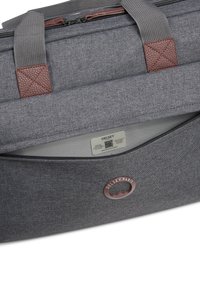 Bolso de tela gris de Delsey Paris con detalles en cuero marrón, bolsillo con cremallera y logo de la marca visible cerca de la abertura del bolsillo.
