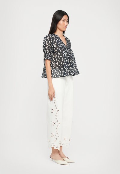 Schwarze, transparente Bluse mit weißem Blumenmuster, kurzen Puffärmeln und V-Ausschnitt. Kombiniert mit weißen, weit geschnittenen Hosen mit bestickten Akzenten.
