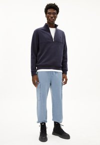 Sweatshirt bleu marine à col zippé avec un t-shirt blanc, associé à un pantalon cargo bleu clair avec des poches latérales et des bottes noires à lacets.