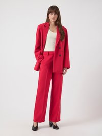 Costume rouge sur mesure avec un blazer croisé et un pantalon à jambes larges. Associé à des chaussures noires à talons hauts. Tissu lisse, accents minimaux.