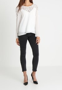 Camicia bianca con sovrapposizione di pizzo sulle spalle, maniche lunghe; abbinata a jeans neri slim-fit con zip alla caviglia e tacchi alti neri.