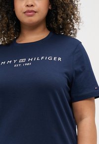 Tumši zila kokvilnas T-krekls ar baltu logo tekstu "TOMMY HILFIGER EST. 1985" uz priekšpuses un nelielu logo akcentu uz piedurknes.
