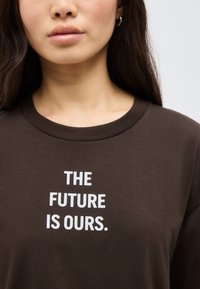 Γυναίκα που φοράει σκουρό καφέ πουκάμισο με λευκό κείμενο «ΤΟ ΜΕΛΛΟΝ ΕΙΝΑΙ ΔΙΚΟ ΜΑΣ» στο κέντρο του στήθους, ορατή από τους ώμους μέχρι τα χείλη.