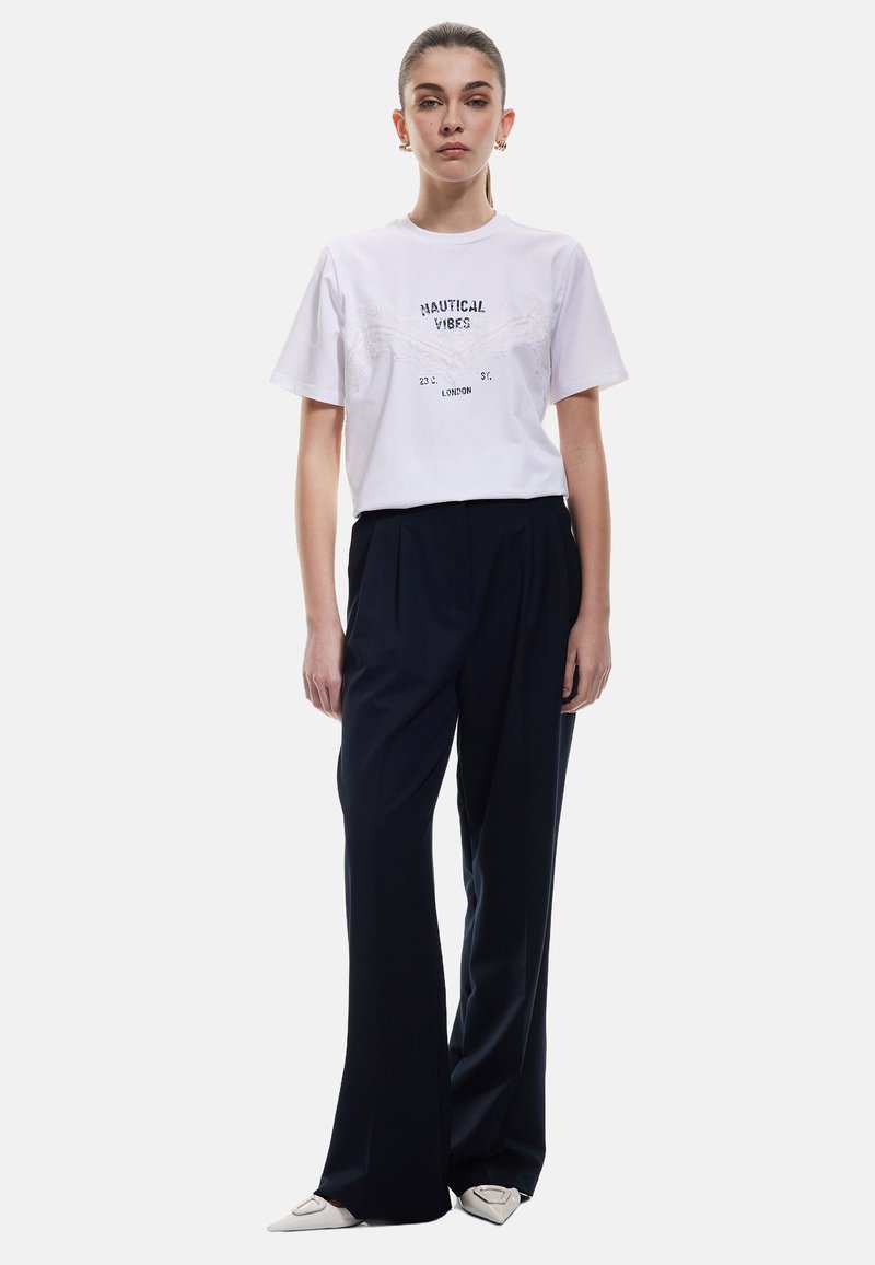 Hvid t-shirt med "NAUTICAL VIBES" tryk, parret med højtaljede, vide navy bukser. Skoene har en spids tå-design.