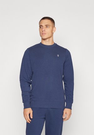 Mand stående iført matchende mørkeblå sweatshirt og bukser, kigger let til højre mod en ensfarvet lys baggrund.