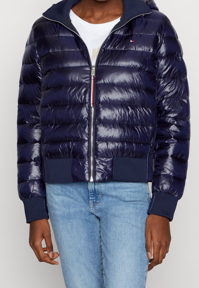 Granatowa pikowana kurtka puchowa z zamkiem błyskawicznym, ściągaczami na rękawach i dole, z połyskującymi przeszyciami i kontrastowym kapturem. Noszona na białej koszuli.