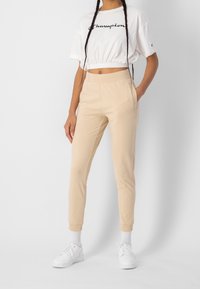 Beige Jogginghosen mit elastischen Bündchen, kombiniert mit einem weißen, kurz geschnittenen T-Shirt mit schwarzem "Champion"-Logo und geflochtenen Haaren. Weiße Turnschuhe.