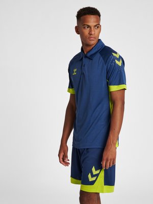 Junger Mann trägt ein Sport-Polo in Navyblau und Limettengrün und passende Shorts, steht mit leicht seitlich gedrehtem Kopf.