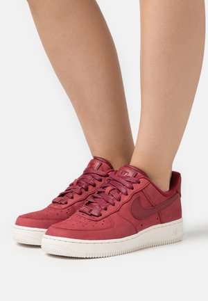 Jambes portant des baskets basses Nike rouges avec des semelles blanches, debout sur une surface blanche.
