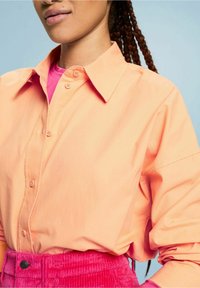 Femme portant une chemise pêche boutonnée sur un top rose avec un pantalon en velours côtelé rose vif, sur fond bleu clair.