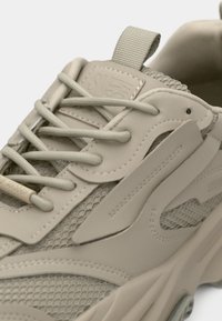 Beige athletic sneaker featuring a mesh upper, textured accents, and flat laces. Side logo and pull tab enhance functionality and design.