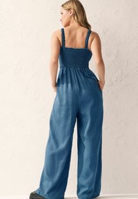 Farmer overall simlé bodrral, széles lábakkal és állítható pántokkal, egységes középkék színben. Texturált anyag, amely jól simul.