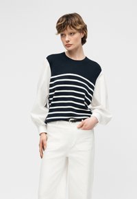 Jeune personne aux cheveux courts portant un gilet sans manches bleu marine à rayures horizontales blanches sur un chemisier blanc à manches longues et un pantalon blanc.