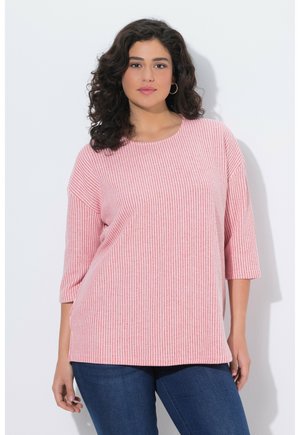 Mujer con cabello rizado oscuro, que lleva pendientes de aro, un suéter suelto a rayas rosas y blancas, y jeans azules, de pie contra un fondo blanco.