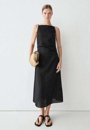 Frau trägt ein ärmelloses schwarzes Midikleid mit dünnen Trägern, hält eine beige geflochtene Clutch und trägt schwarze Sandalen, steht vor einer schlichten Wand.