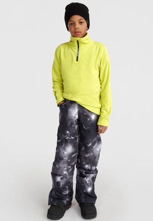 Un pull en molleton jaune vif avec un col montant, associé à un pantalon de neige noir et gris à motifs. Un bonnet noir et des bottes noires robustes.