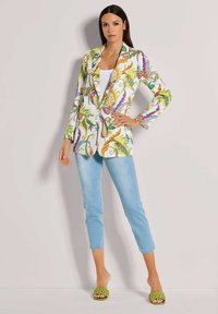 Frau steht gegen eine beige Wand und trägt ein farbenfrohes Blumensakko, ein weißes Oberteil, helle, cropped Jeans und grüne, geflochtene Open-Toe-Absätze.