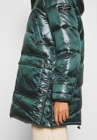 Grön puffjacka med en glänsande yta, med dragkedja framtill och sidofickor, framträdande sömmar och en logotag på axeln.