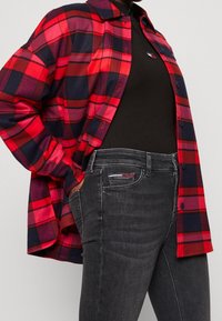 Camicia di flanella a quadri rossi e blu con bottoni e taschino sul petto, indossata sopra una maglietta nera, abbinata a jeans grigio scuro.