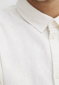 Chemise en lin beige clair avec col, fermeture à boutons et tissu texturé présentant un motif de tissage subtil. Matière douce et respirante.