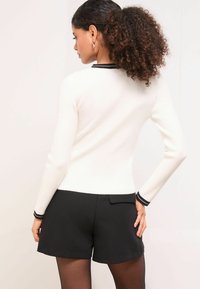 Fehér bordázott pulóver fekete szegéllyel, testhezálló kialakítás, hosszú ujjú és kerek nyakkivágással, magas derekú fekete rövidnadrággal párosítva.