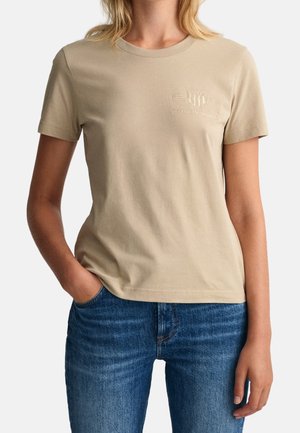 Kobieta ubrane w beżowy t-shirt z krótkim rękawem z subtelnym logo na piersi i niebieskie dżinsy, jedna ręka w kieszeni, na jednolitym tle.