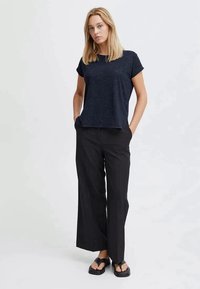 Top blu scuro a maniche corte realizzato in tessuto testurizzato, abbinato a pantaloni neri a gamba larga. La persona in posa ha le mani nelle tasche, indossando sandali neri.