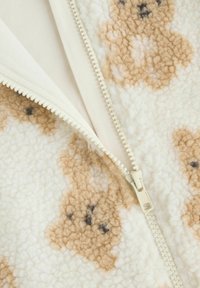 Fleece-Jacke mit cremiger Basis, ausgestattet mit einem braunen Bärenmuster und schwarzen Akzenten. Reißverschluss mit cremigem Besatz und weicher Textur.