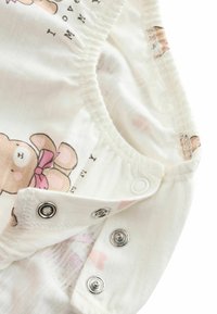 Combinaison en tissu blanc avec un motif d'ours et un nœud rose. Boutons-pression en bas et texture douce et extensible. Des motifs de cœurs subtils sont visibles.