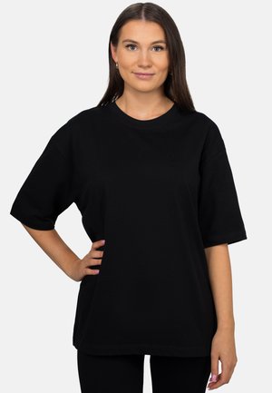 Junge Frau mit langen braunen Haaren steht frontal, trägt ein schlichtes schwarzes übergroßes T-Shirt und hat eine Hand in der Hüfte vor weißem Hintergrund.