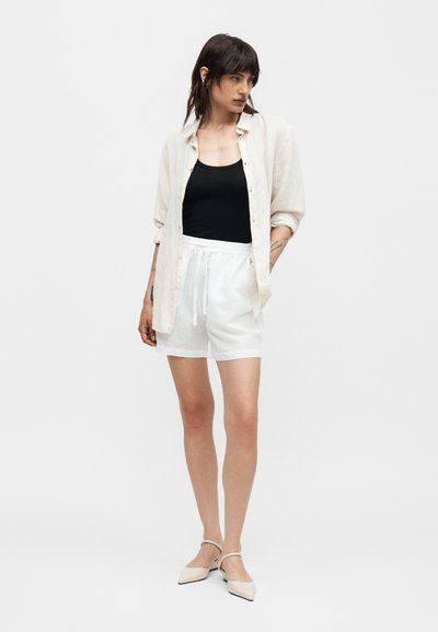 Γυναίκα στέκεται φορώντας μαύρο αμάνικο μπλουζάκι, χαλαρό λευκό πουκάμισο με κουμπιά, λευκά σορτς με κορδόνι και μπεζ μυτερές μπαλαρίνες με λουράκια στον αστράγαλο.