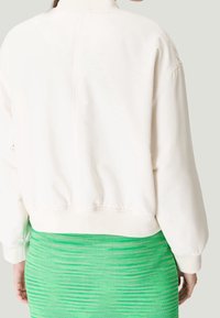 Pull-over couleur crème avec ourlet et poignets côtelés, présentant un design minimaliste et une texture douce. Porté avec une jupe verte à motifs.