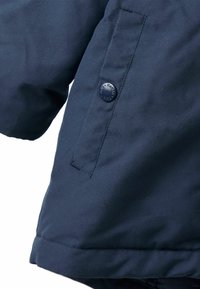 Jūras zilas izolētas jakas detaļas, kuras izceļ gluda auduma tekstūra, izteiksmīga pogas ar iepresētu logo un sānu šuvē ievietota kabata.