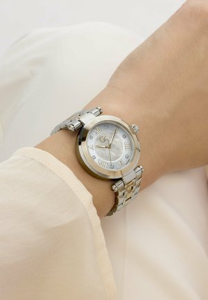 Sidabrinis riešinis laikrodis su romėniškomis skaitmenimis ir deimanto formos valandų žymekliais ant kairiojo žmogaus riešo, dėvinčio šviesiai smėlinės spalvos ilgomis rankovėmis marškinius.