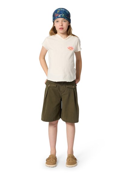 Enfant portant un t-shirt blanc, un short olive jusqu'aux genoux, des chaussures à enfiler couleur beige et un foulard bleu à fleurs, debout les mains dans les poches.