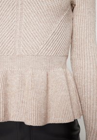 Maglione lavorato a maglia di un beige chiaro con una texture a coste. Presenta un orlo a peplum e un motivo diagonale nella parte superiore. Include maniche lunghe.