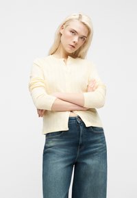 Νέα γυναίκα με μακριά ξανθά μαλλιά, που φοράει ανοιχτόκίτρινη ζακέτα και μπλε τζιν, στέκεται με σταυρωμένα χέρια μπροστά σε απλό φόντο.