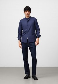 Camicia blu navy con colletto alla mandarina, abbinata a jeans blu scuro e scarpe nere, che mette in evidenza una texture di tessuto liscia.