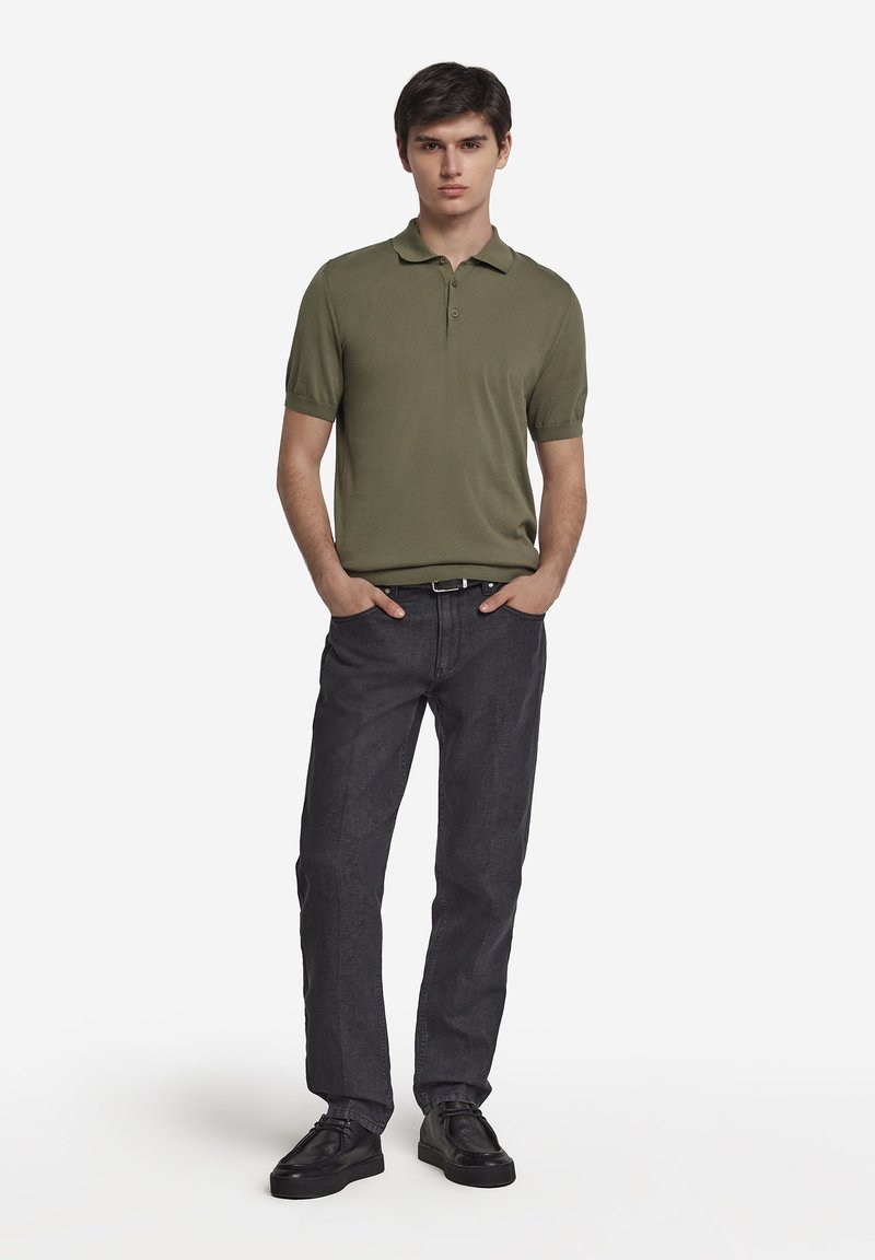 Olivgrön polo t-shirt med korta ärmar, kombinerad med mörkgrå jeans och svarta snörskor, bärs av en man som står med händerna i fickorna.